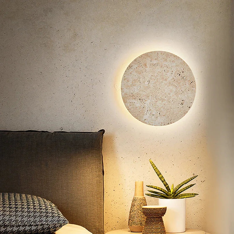 Stone disc wall light with a warm halo above a bed with a plant on the nightstand
